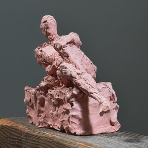 May include: A pink clay sculpture of a seated figure, with rough, textured surfaces. The figure is posed in a relaxed position, with one arm resting on the knee. The sculpture sits on a wooden surface against a gray background.