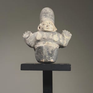 Puede incluir: Una figurita de cerámica gris de una persona que lleva un sombrero alto y un cinturón decorativo. La figurita tiene los brazos levantados y está de pie sobre un soporte negro.