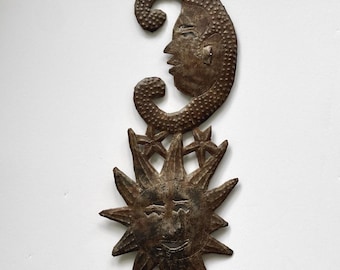 May include: A rustic metal wall hanging featuring a sun and moon design. The sun has a smiling face with pointed rays, and the moon has a detailed face. The piece is a dark brown colour and includes stars.