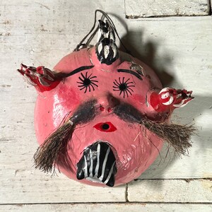 Puede incluir: Una m&aacute;scara de papel mach&eacute; rosa con detalles pintados en negro y blanco, incluyendo ojos, bigote y un cuerno. La m&aacute;scara tiene acentos pintados en rojo y blanco en los lados y un peque&ntilde;o agujero en el centro de la boca.