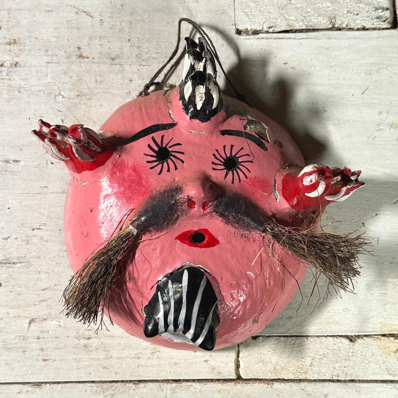 Puede incluir: Una m&aacute;scara de papel mach&eacute; rosa con detalles pintados en negro y blanco, incluyendo ojos, bigote y un cuerno. La m&aacute;scara tiene acentos pintados en rojo y blanco en los lados y un peque&ntilde;o agujero en el centro de la boca.