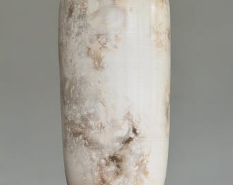 May include: A tall, cylindrical ceramic vase with a rounded top. The vase is primarily off-white with mottled brown and tan markings. It stands on a wooden surface against a gray backdrop. The vase has a smooth, glossy finish.