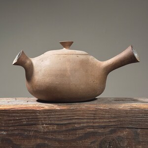May include: A brown ceramic teapot with a rounded body and a spout on the side. The teapot is sitting on a wooden surface.