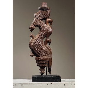 May include: A dark brown wooden sculpture of a mythical creature with intricate details. The sculpture is mounted on a black wooden base.