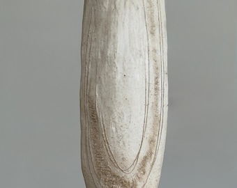 May include: A tall, off-white ceramic vase with a textured surface and vertical lines. The vase has a rounded top and a slightly wider base. The design includes concentric, curved lines near the center. The vase is displayed on a wooden surface.