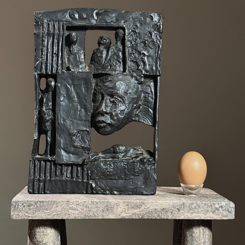 May include: A black abstract sculpture with a human face and figures in relief. The sculpture is on a stone pedestal with a brown egg in a small white bowl.