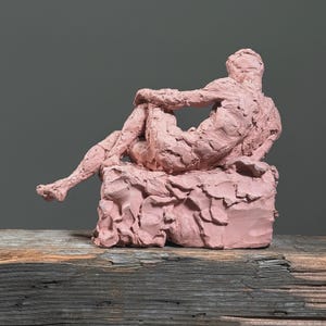 May include: A pink clay sculpture of a reclining figure. The figure is seated on a textured, rectangular base, with one leg extended and arms crossed. The sculpture has a rough, textured surface and is set against a gray background.