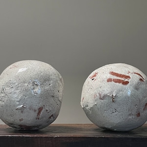 May include: Two spherical ceramic sculptures with a cracked, off-white glaze and reddish-brown accents. The spheres are displayed on a dark wooden surface against a neutral background. The texture and color variations give them a rustic appearance.