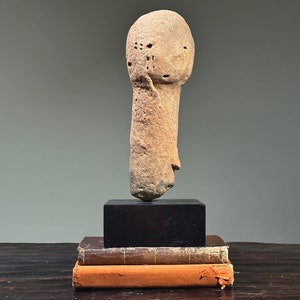 May include: A terracotta sculpture of a human head and neck, with a rough, textured surface. The sculpture is displayed on a black wooden base and a stack of books.