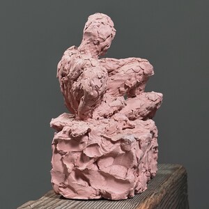 May include: A pink clay sculpture of a seated figure, with a rough, textured surface. The figure is positioned on a textured base, with the back of the figure facing the viewer. The sculpture is set against a gray background.
