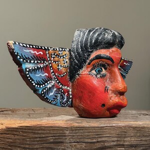 May include: A hand-carved wooden mask with a red face, black hair, and blue eyes. The mask has decorative wings painted with blue, red, and orange patterns. The mask is displayed on a wooden surface.