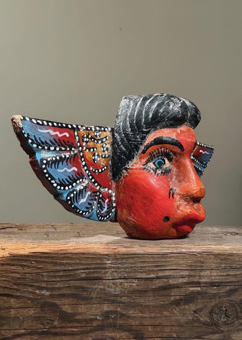 May include: A hand-carved wooden mask with a red face, black hair, and blue eyes. The mask has decorative wings painted with blue, red, and orange patterns. The mask is displayed on a wooden surface.