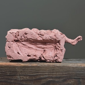 May include: A textured, pink sculpture with an abstract form, possibly clay or a similar material. The sculpture has a rough, uneven surface and sits on a wooden surface. The background is a solid gray.