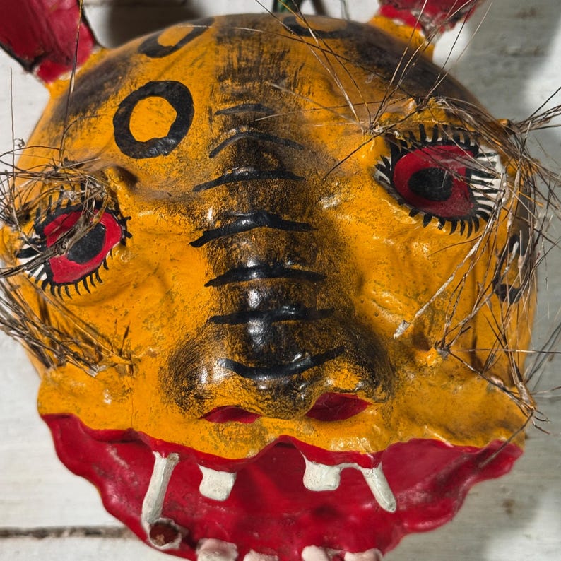 Puede incluir: Una m&aacute;scara de papel mach&eacute; amarilla y roja con detalles pintados en negro y largos pelos negros. La m&aacute;scara tiene una gran sonrisa con dientes y ojos rojos.