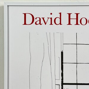 May include: A white framed print with the text "David Hockney" in red lettering. The print features a black and white line drawing of a window with a grid pattern.