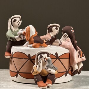 May include: A handcrafted ceramic sculpture featuring figures in traditional attire. The figures are arranged around a circular base with geometric patterns in brown and white. One figure holds a bird. The sculpture has a matte finish.