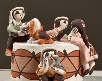May include: A handcrafted ceramic sculpture featuring figures in traditional attire. The figures are arranged around a circular base with geometric patterns in brown and white. One figure holds a bird. The sculpture has a matte finish.