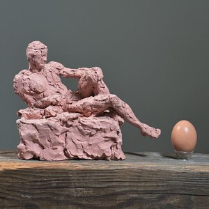 May include: A pink clay sculpture of a reclining figure on a textured base. A brown egg sits on a clear stand to the right of the sculpture. The sculpture and egg are on a wooden surface.