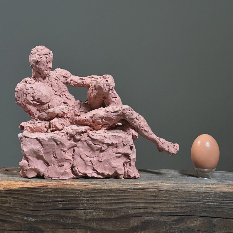 May include: A pink clay sculpture of a reclining figure on a textured base. A brown egg sits on a clear stand to the right of the sculpture. The sculpture and egg are on a wooden surface.