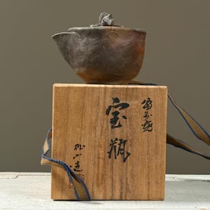 Può includere: Una piccola teiera in ceramica marrone scuro con coperchio, appoggiata su una scatola di legno. La scatola ha caratteri giapponesi stampati. Un nastro blu e oro è attaccato alla scatola.