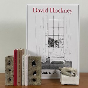 May include: A set of four books with white covers and black text titles. The books are stacked on top of each other and are being held up by two gray concrete blocks. The books are titled "The Freedom Within Us", "Kara Walker, My Complement, My Lacно. Му Оррегльок, My Love", "DR. IKKAKU OCHI COLLECTION", and "The Diary of a Nose JEAN-CLAUDE ELLENA". Behind the books is a white poster with black line drawings of a window, curtains, and a chair. The poster has the text "David Hockney" at the top and "SIANA-7/2-7" at the bottom.