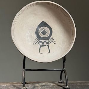May include: A white ceramic bowl with a black and white spider design on a black metal stand. The bowl is on a wooden surface.