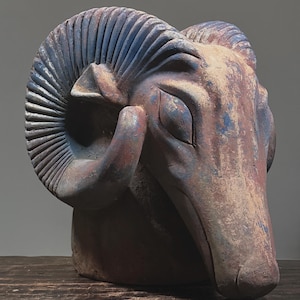 May include: A weathered, ceramic ram head sculpture. The sculpture features large, spiraling horns and is painted in shades of brown, blue, and red. The ram's face has closed eyes and a detailed texture, suggesting an aged appearance.