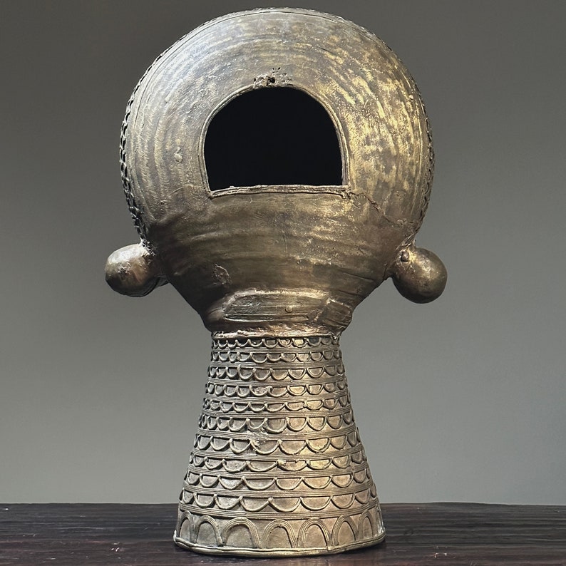 May include: A gold-colored ceramic sculpture of a head with a round, hollow top and a decorative base. The head has a large, circular opening in the front.