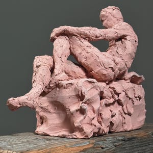 May include: A pink clay sculpture of a seated figure, with arms wrapped around the knees. The figure is sitting on a textured, block-like base. The sculpture is displayed on a wooden surface against a gray background.