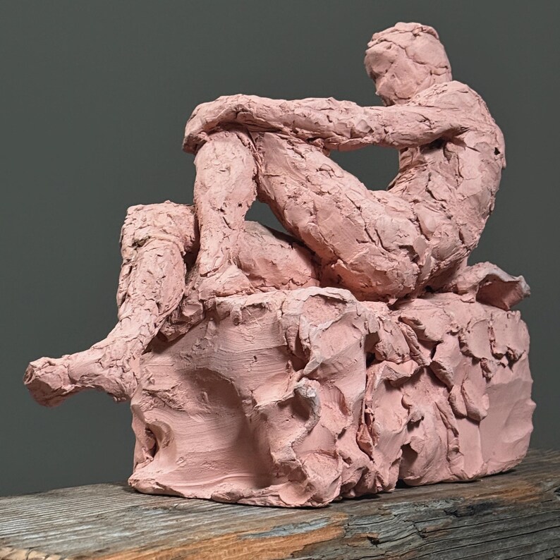 May include: A pink clay sculpture of a seated figure, with arms wrapped around the knees. The figure is sitting on a textured, block-like base. The sculpture is displayed on a wooden surface against a gray background.