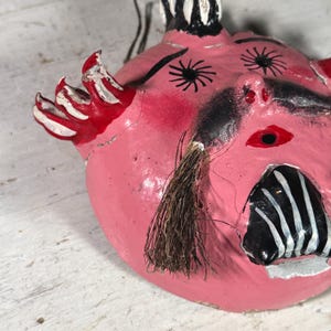 Puede incluir: Una m&aacute;scara de papel mach&eacute; rosa con detalles pintados en negro y blanco. La m&aacute;scara tiene una boca grande y exagerada con rayas negras y blancas, y un largo bigote negro con un mech&oacute;n de pelo marr&oacute;n. Los ojos son grandes y negros con estrellas blancas.