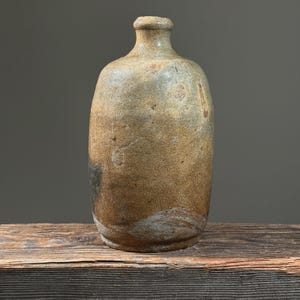 May include: A rustic, handcrafted stoneware bottle with a narrow neck and rounded body. The bottle has a mottled, earthy color palette of browns, tans, and hints of gray, with a textured surface. The bottle is displayed on a wooden surface.