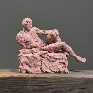 May include: A pink sculpture of a reclining figure, resting on a textured base. The figure is in a relaxed pose, with one arm bent and the other resting on the base. The sculpture is made of a rough, textured material.