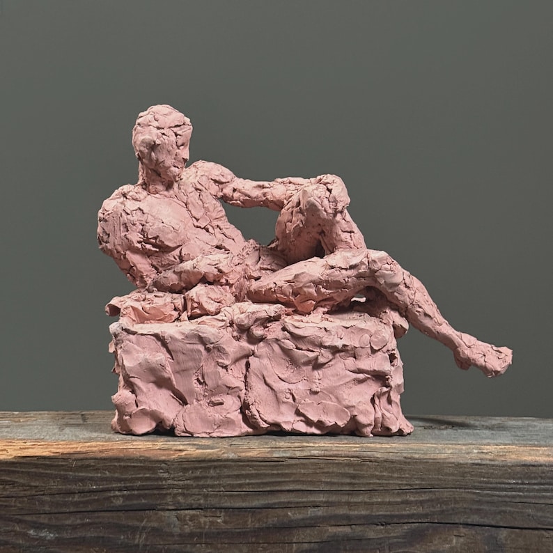 May include: A pink sculpture of a reclining figure, resting on a textured base. The figure is in a relaxed pose, with one arm bent and the other resting on the base. The sculpture is made of a rough, textured material.
