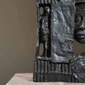 May include: Black abstract sculpture with a face and a figure carved into the side. The sculpture is made of a rough, textured material.