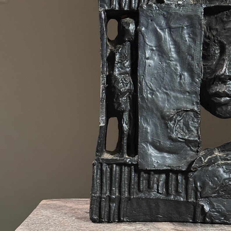 May include: Black abstract sculpture with a face and a figure carved into the side. The sculpture is made of a rough, textured material.