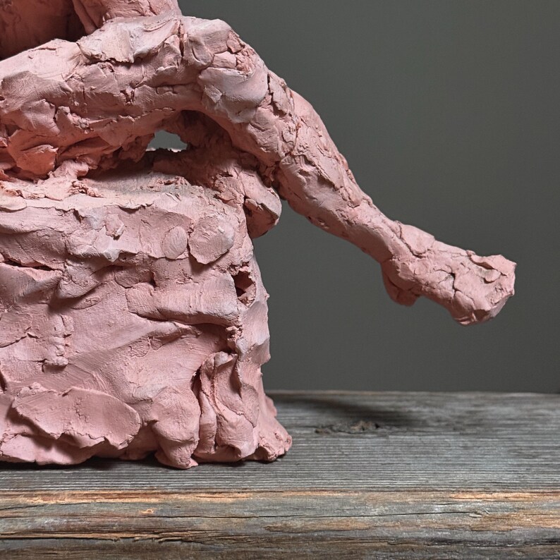 May include: A pink clay sculpture of a seated figure, with an arm extended. The sculpture has a rough, textured surface and is set against a neutral gray background. The figure appears to be in a relaxed pose.