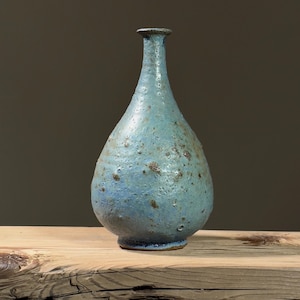 Puede incluir: Un jarrón de cerámica azul con una superficie texturizada y un cuerpo redondeado. El jarrón tiene un cuello estrecho y una pequeña abertura.