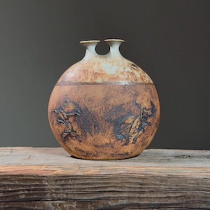 Pode incluir: Um vaso de cerâmica marrom e branco com duas aberturas. O vaso tem uma forma arredondada e é decorado com um esmalte marrom e um esmalte branco. O vaso está sobre uma superfície de madeira.