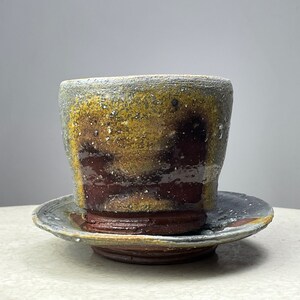 Puede incluir: Un juego de taza y platillo de cerámica con un esmalte marrón, amarillo y gris. La taza y el platillo tienen un aspecto rústico y hecho a mano.