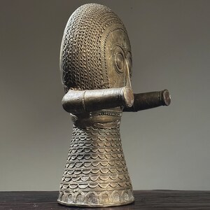 May include: A gold-colored metal sculpture of a stylized human figure with a patterned head and a long, cylindrical arm. The sculpture has a textured surface and a rounded base.