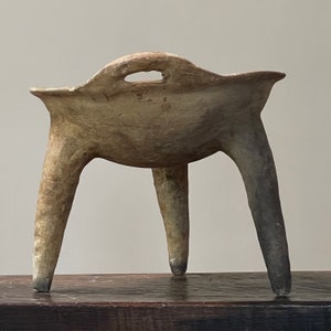 May include: A three-legged ceramic bowl with a handle. The bowl is a light brown color and has a rough, textured surface.