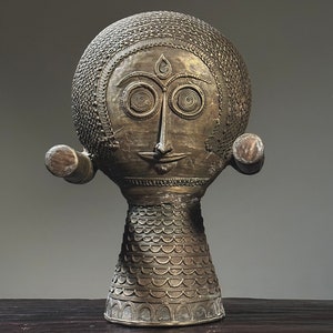 May include: A gold-colored metal sculpture of a stylized human head with intricate details. The head has a round shape with a textured surface and a face with large eyes, a small nose, and a slightly smiling mouth. The sculpture is mounted on a base with a textured surface.