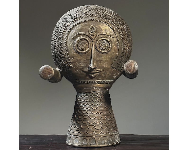 May include: A gold-colored metal sculpture of a stylized human head with intricate details. The head has a round shape with a textured surface and a face with large eyes, a small nose, and a slightly smiling mouth. The sculpture is mounted on a base with a textured surface.