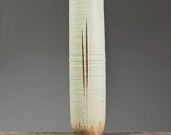 May include: A tall, cylindrical ceramic vase with a cream-colored glaze and a vertical split down the front. The vase has a textured surface with horizontal ridges and a slightly flared rim. The base and rim have a warm, earthy tone.