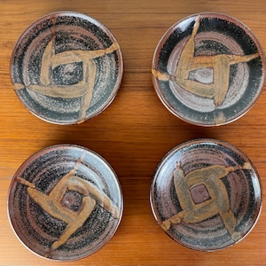 May include: Set of four small ceramic plates with a black, brown, and tan swirl design. Each plate features a stylized, abstract design in the center.