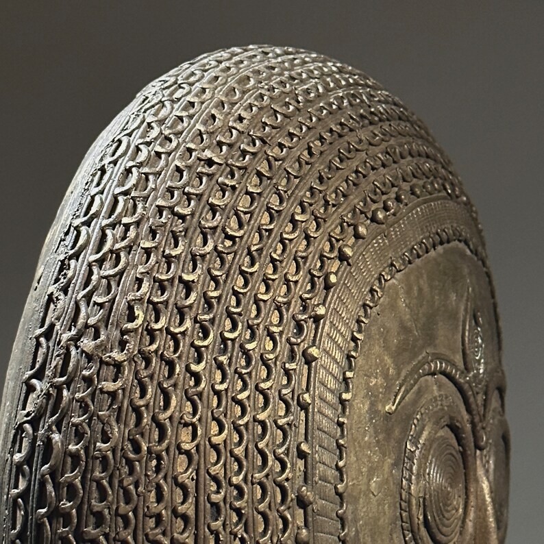 May include: A bronze sculpture of a head with intricate details. The head is covered in a pattern of small, raised circles and lines. The sculpture is a traditional African art piece.