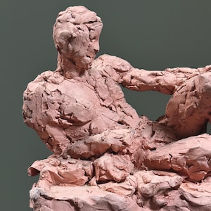May include: A reddish-brown clay sculpture of a reclining figure. The figure is in a relaxed pose, with one arm extended and the other bent. The sculpture has a rough, textured surface and is set against a muted green background.