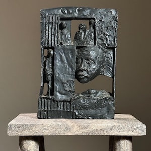 May include: A black abstract sculpture with a face and figures carved into it. The sculpture is made of a rough, textured material and has a geometric design.