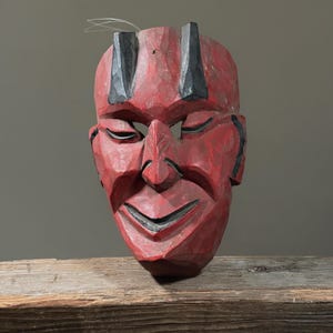 Puede incluir: Una máscara de madera tallada, principalmente roja con detalles negros. La máscara presenta una boca sonriente, ojos cerrados y elementos puntiagudos. Un alambre está sujeto en la parte superior. Descansa sobre una superficie de madera.